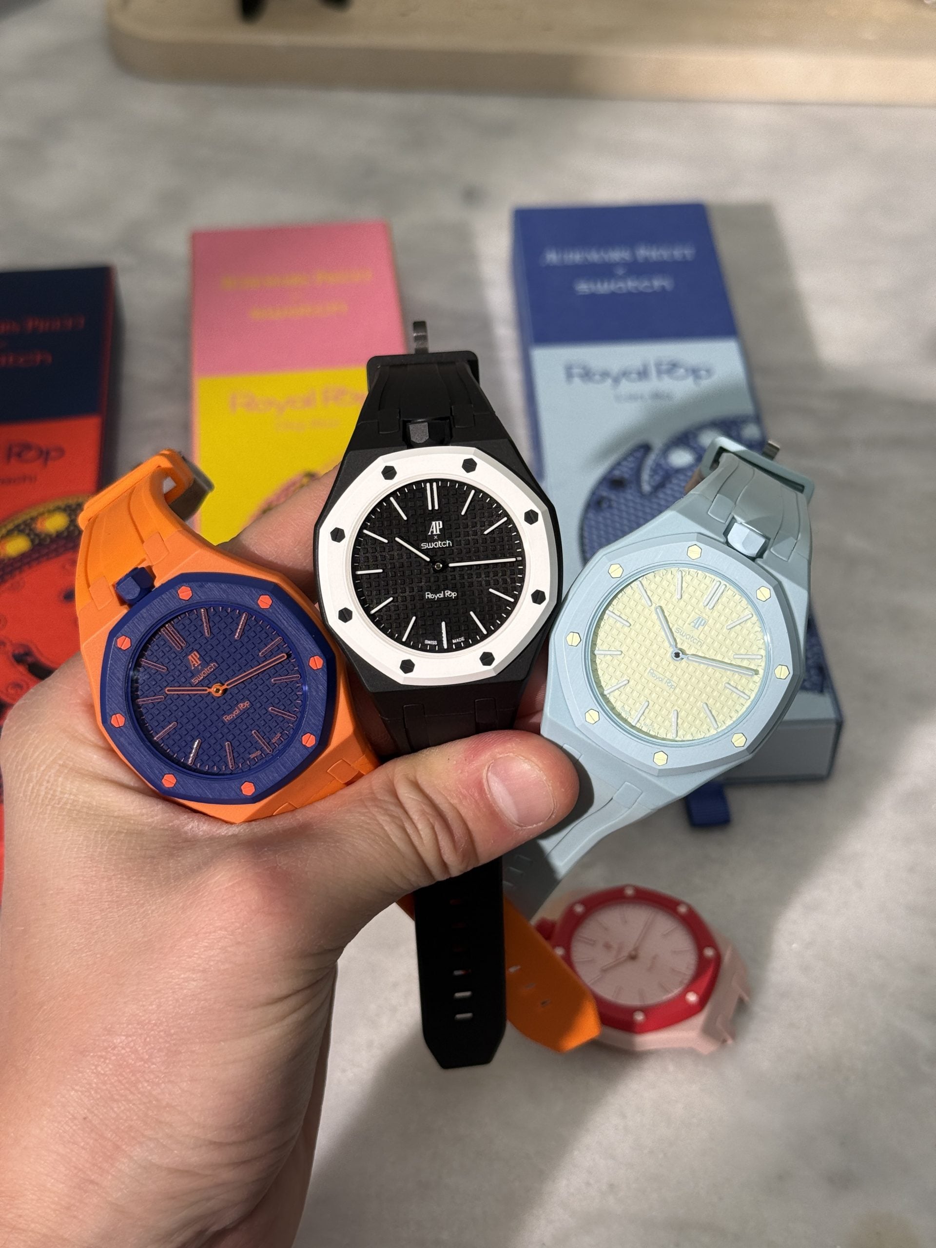 Tre AP × Swatch Royal Pop med monterade straps hållna i handen – Blaue Acht på grå rem, Ocho Negro på svart rem och Orenji Hachi på orange rem, hands-on