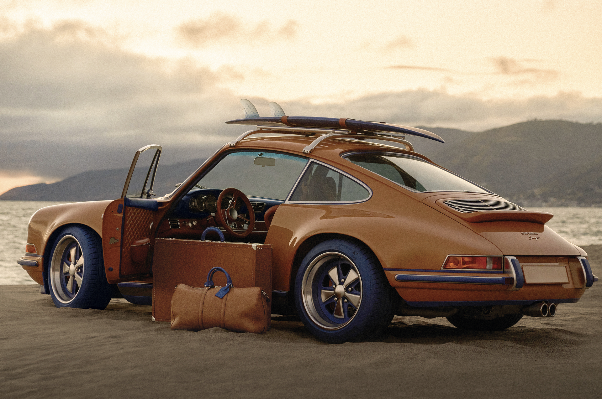 porsche 911 reimagined by singer i saffransgult med koboltblå fälgdetaljer och surfbrädor på takräcket vid stranden