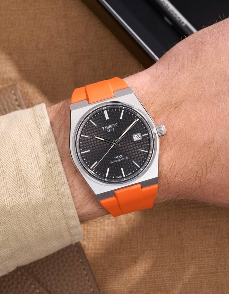 tissot prx med svart urtavla och orange gummiarmband på handleden under en manschett