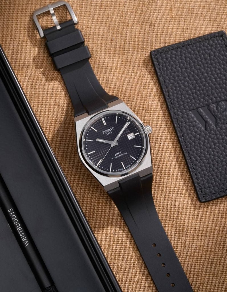 tissot prx med svart urtavla och svart gummiarmband bredvid ett läderfodral
