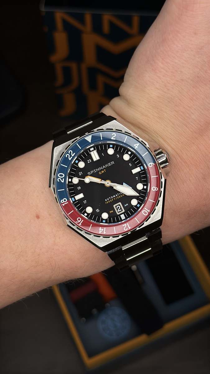 närbild på spinnaker dumas gmt automatic på handleden med svart urtavla, orange gmt-visare och datumfönster vid 4:30